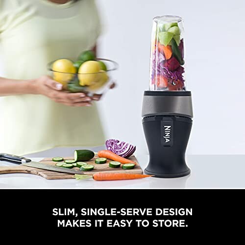 Person holding a bowl of fruit next to a Ninja blender with vegetables inside.